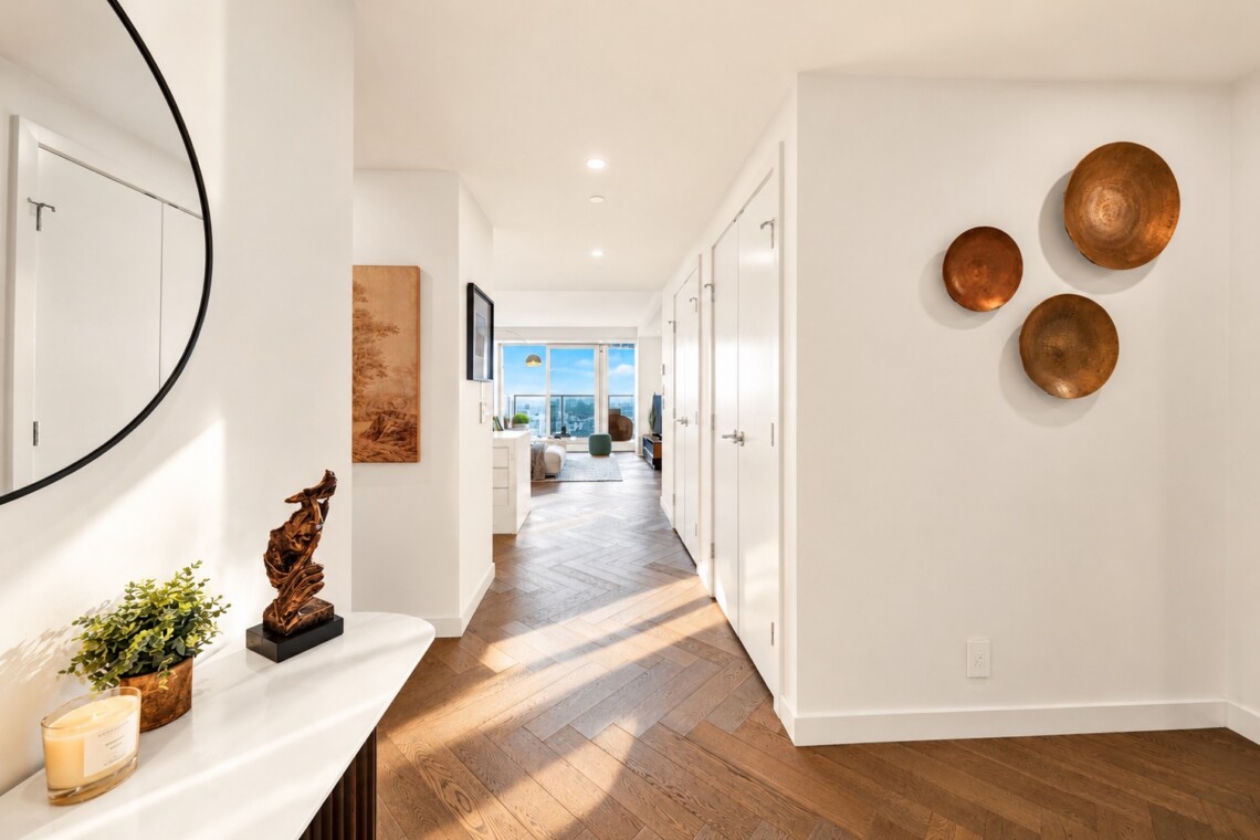 Bright white hallway with a round wall mirror on the left, a console table carrying a plant and a wooden sculpture, and copper plates on the right wall; opens to a living area with a city view.