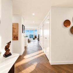 Bright white hallway with a round wall mirror on the left, a console table carrying a plant and a wooden sculpture, and copper plates on the right wall; opens to a living area with a city view.