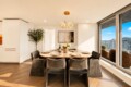 Round marble dining table set for eight with grey upholstered chairs in a bright modern dining room; chandelier overhead and city views through floor-to-ceiling windows.