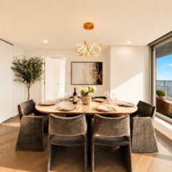 Round marble dining table set for eight with grey upholstered chairs in a bright modern dining room; chandelier overhead and city views through floor-to-ceiling windows.