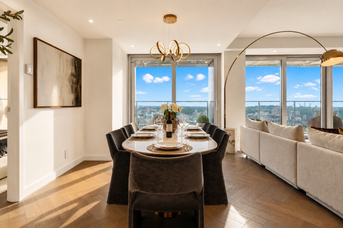 Bright open dining area with a long oval table set for eight, wine bottle and white flower centerpiece, and large windows with a city view.