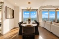 Bright open dining area with a long oval table set for eight, wine bottle and white flower centerpiece, and large windows with a city view.