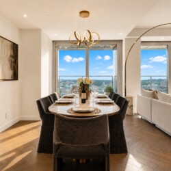 Bright open dining area with a long oval table set for eight, wine bottle and white flower centerpiece, and large windows with a city view.