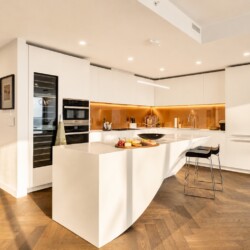 Modern white kitchen with a curved island, warm golden backsplash, and black bar stools.
