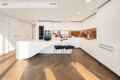 Modern white kitchen with curved island, copper backsplash, and two black bar stools on herringbone wood floor.