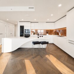 Modern white kitchen with curved island, copper backsplash, and two black bar stools on herringbone wood floor.