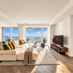 Bright open living room with beige sectional, colorful pillows, and a rug overlooking the city from large windows yes.