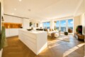 Bright open-plan kitchen and living area with a large white island, white cabinets, and a cozy seating area by expansive windows overlooking the city skyline