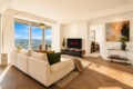 Bright modern living room with a beige sectional sofa facing a TV that shows Netflix, large windows with a city view, and wood floors.