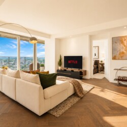 Bright modern living room with a beige sectional sofa facing a TV that shows Netflix, large windows with a city view, and wood floors.