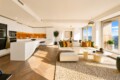 Open-concept living room and kitchen with white cabinetry, warm orange backsplash, and a beige sectional; large windows overlook a city view.