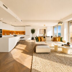 Open-concept living room and kitchen with white cabinetry, warm orange backsplash, and a beige sectional; large windows overlook a city view.