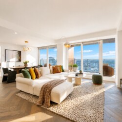 Bright modern living room with a white sectional, colorful cushions, and large windows overlooking the city skyline.