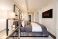 Contemporary hotel bedroom with a plush gray upholstered bed, abstract art above, and a wall-mounted Netflix screen on the right side.