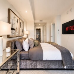 Contemporary hotel bedroom with a plush gray upholstered bed, abstract art above, and a wall-mounted Netflix screen on the right side.