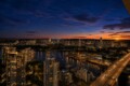 Aerial view of a city at dusk: river with boats, marina, high-rise buildings, and a lit bridge on the right edge.