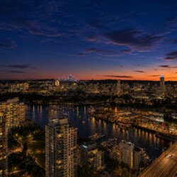 Aerial view of a city at dusk: river with boats, marina, high-rise buildings, and a lit bridge on the right edge.
