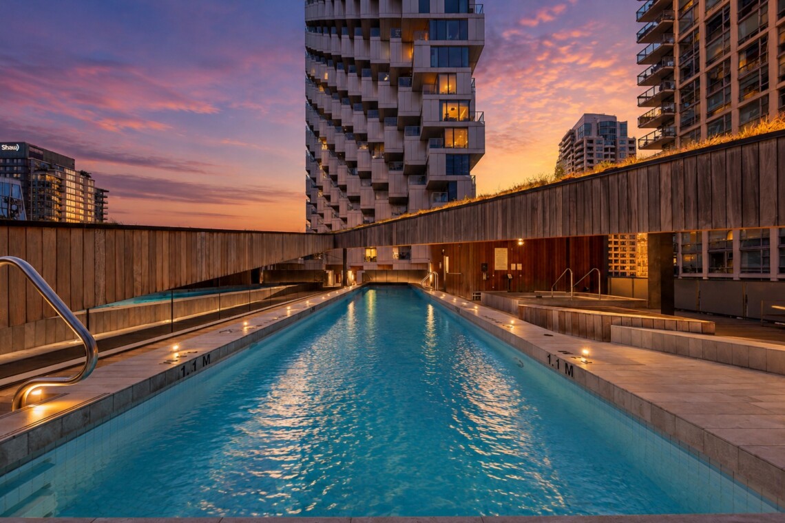 Rooftop swimming pool at sunset, surrounded by wooden walls and modern high-rise buildings. The sky turns orange-pink above a unique, stacked apartment tower.
