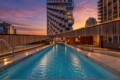 Rooftop swimming pool at sunset, surrounded by wooden walls and modern high-rise buildings. The sky turns orange-pink above a unique, stacked apartment tower.