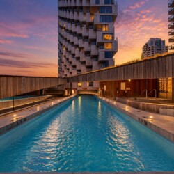 Rooftop swimming pool at sunset, surrounded by wooden walls and modern high-rise buildings. The sky turns orange-pink above a unique, stacked apartment tower.
