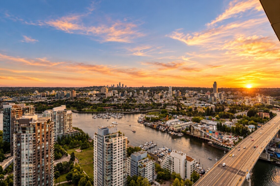 Aerial cityscape at sunset: river with boats, marina, tall buildings along the waterfront, and a busy bridge on the right under a warm orange sky