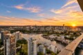 Aerial cityscape at sunset: river with boats, marina, tall buildings along the waterfront, and a busy bridge on the right under a warm orange sky