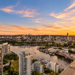 Aerial cityscape at sunset: river with boats, marina, tall buildings along the waterfront, and a busy bridge on the right under a warm orange sky