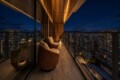 Night view from a high-rise balcony with two wicker lounge chairs and warm lighting over a cityscape at night.