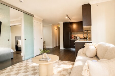 Open-plan living area with a cream sofa, round coffee table, and a dark wood kitchen; frosted glass sliding doors lead to a bedroom.