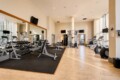 Well-lit gym interior with a mirrored wall, free weights on racks, benches, and weight machines along a black rubber mat area near cardio machines beyond the windows.