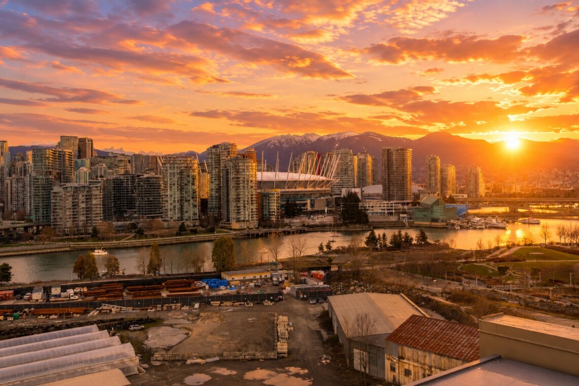 Sunset over a dense city skyline by a river, with tall glass buildings and a distant mountain range bathed in orange light.