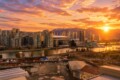 Sunset over a dense city skyline by a river, with tall glass buildings and a distant mountain range bathed in orange light.