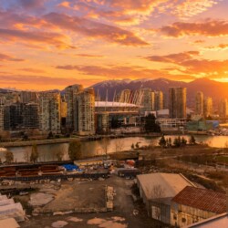 Sunset over a dense city skyline by a river, with tall glass buildings and a distant mountain range bathed in orange light.