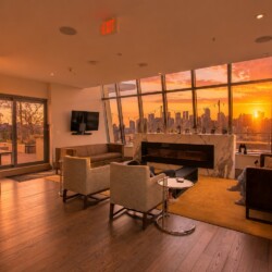 Cozy modern lounge with a marble fireplace, beige chairs, and a city skyline at sunset through large angled windows