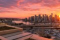 Sunset over a city skyline with tall buildings by a marina, boats in the harbor, and a highway overpass in the foreground.