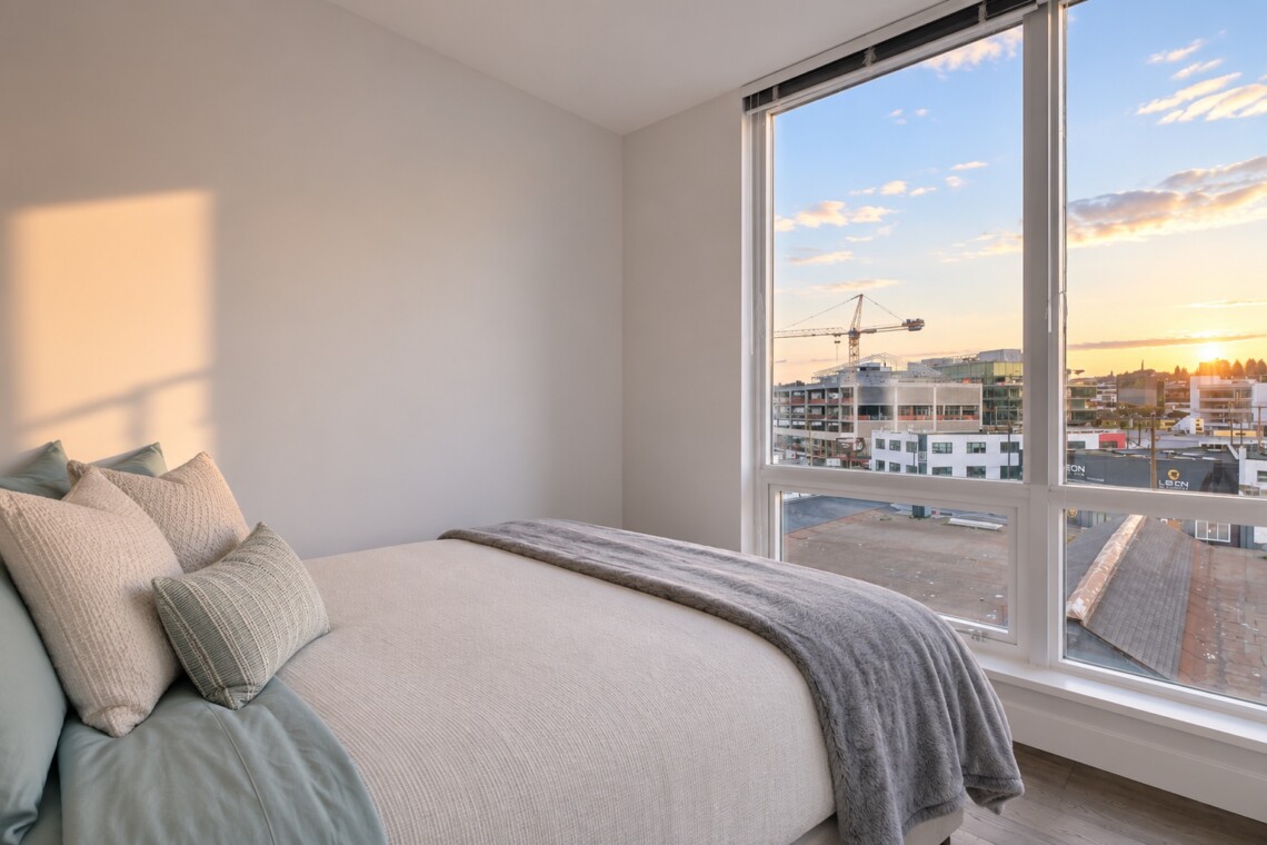 Bedroom with a large window view of a city at sunset; bed dressed in cream linens with pillows and a gray throw.