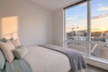 Bedroom with a large window view of a city at sunset; bed dressed in cream linens with pillows and a gray throw.