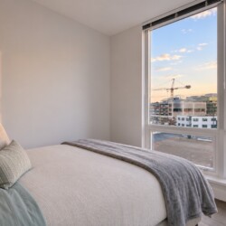 Bedroom with a large window view of a city at sunset; bed dressed in cream linens with pillows and a gray throw.