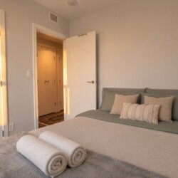 Sunlit bedroom with a neatly made bed, green-gray pillows, and two rolled towels at the foot, warm light streaming from a hallway door.