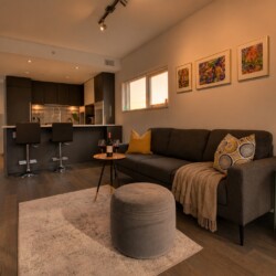 Open-concept living area with a dark kitchen island, two bar stools, gray sofa, and three colorful framed artworks.