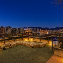 Night view of a city skyline from a rooftop terrace, with a large carved wooden crocodile sculpture in the foreground on tiled ground.