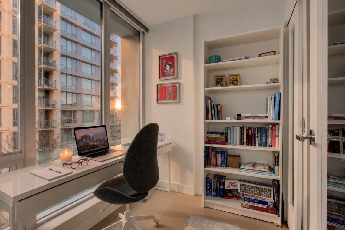 Bright home office with a white desk by a large window, a laptop, glasses, and a lit candle, overlooking a cityscape at sunset.