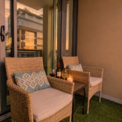 Cozy balcony seating with wicker chairs, cushions, wine bottles, and a lit candle on a small table as evening light shines on neighboring buildings