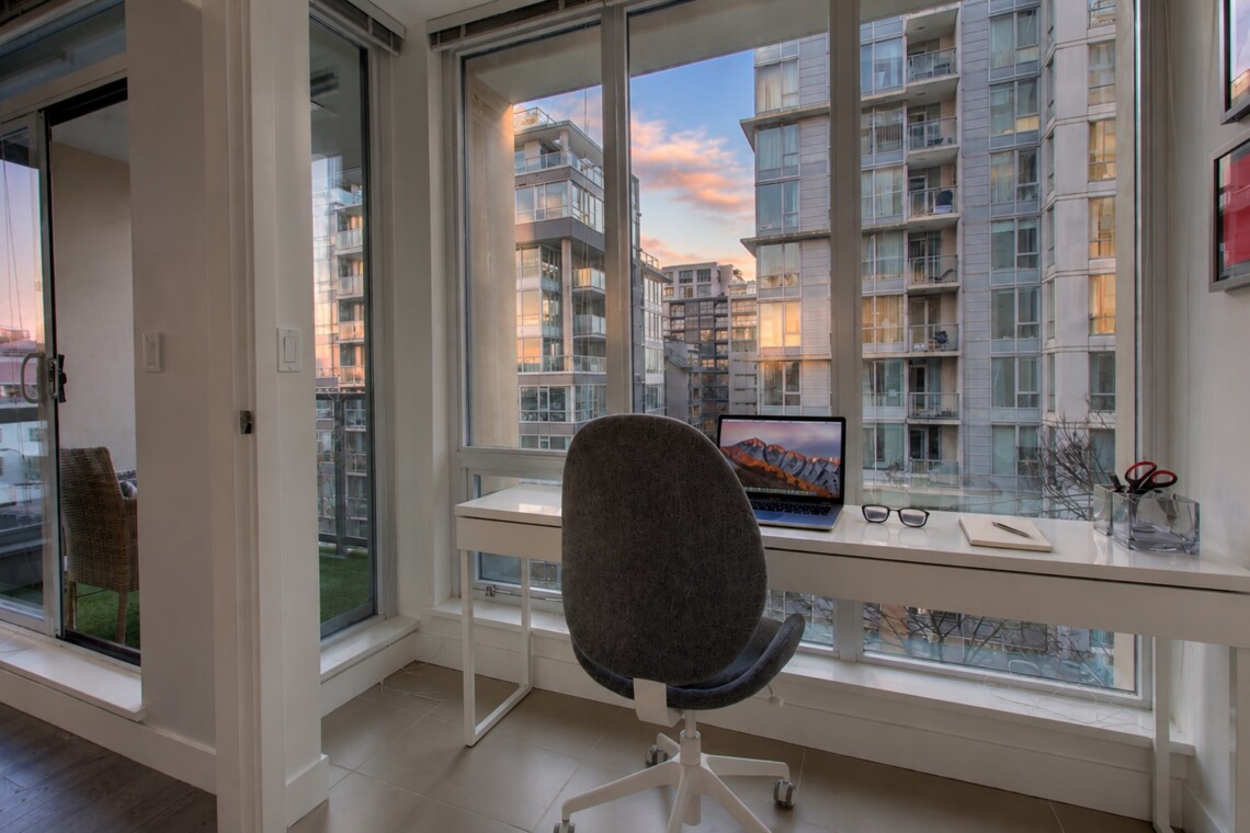 Home office with a white desk by large windows showing a cityscape of modern high-rise buildings at sunset. A laptop, glasses, notebook, and a pen on the desk, with a gray office chair in front.