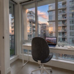 Home office with a white desk by large windows showing a cityscape of modern high-rise buildings at sunset. A laptop, glasses, notebook, and a pen on the desk, with a gray office chair in front.