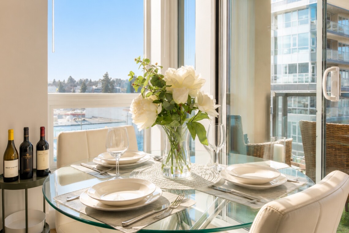 Sunlit dining table set for two with a vase of white flowers and city view through large windows in the background.