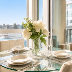 Sunlit dining table set for two with a vase of white flowers and city view through large windows in the background.