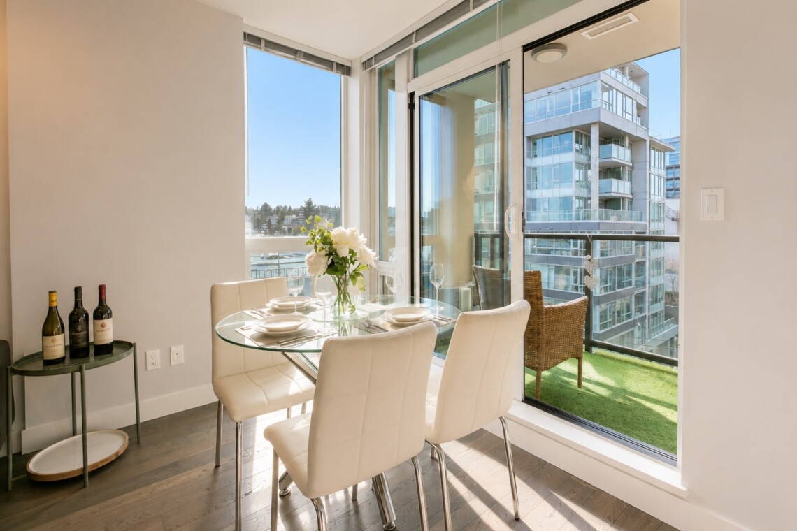 Bright dining area with a glass round table, four white upholstered chairs, and a vase of white flowers by large windows.