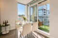 Bright dining area with a glass round table, four white upholstered chairs, and a vase of white flowers by large windows.