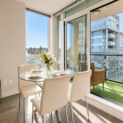 Bright dining area with a glass round table, four white upholstered chairs, and a vase of white flowers by large windows.